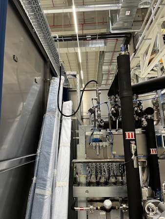 An industrial setting with machinery, visible ductwork, and metal equipment. Pipes and cables are interconnected, and some components are wrapped in protective material. The ceiling features a grid of lights and HVAC ducts.
