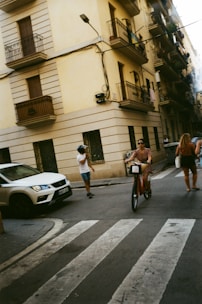 A person is cycling a red rental bike across a street at an intersection with a pedestrian crossing. A white SUV is parked nearby. Two people are walking on the sidewalk, one wearing a white shirt and denim shorts, and the other with a backpack. The background shows a tall building with multiple balconies and closed shutters on the windows.