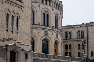 An architectural structure featuring ornate brickwork and arched windows. The facade exhibits intricate masonry with decorative geometric patterns. A railing runs alongside a walkway, leading to an archway with elaborate woodwork. Street lamps are visible, indicating an outdoor setting.