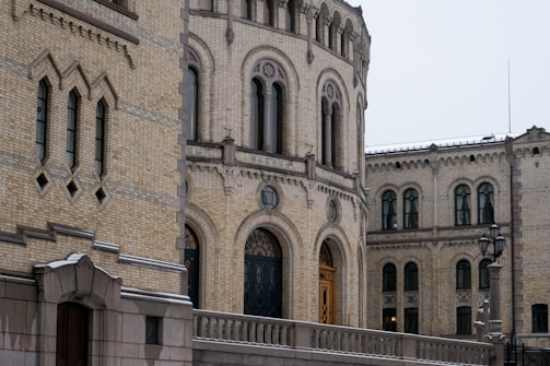 An architectural structure featuring ornate brickwork and arched windows. The facade exhibits intricate masonry with decorative geometric patterns. A railing runs alongside a walkway, leading to an archway with elaborate woodwork. Street lamps are visible, indicating an outdoor setting.
