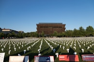 A large field with numerous white flower arrangements placed in rows, each symbolizing a purpose. In the background is a distinctive, modern brown building surrounded by lush trees. Information boards in the foreground provide details about a nonprofit organization and convey messages related to gun violence prevention.
