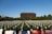 A large field with numerous white flower arrangements placed in rows, each symbolizing a purpose. In the background is a distinctive, modern brown building surrounded by lush trees. Information boards in the foreground provide details about a nonprofit organization and convey messages related to gun violence prevention.