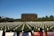 A large field with numerous white flower arrangements placed in rows, each symbolizing a purpose. In the background is a distinctive, modern brown building surrounded by lush trees. Information boards in the foreground provide details about a nonprofit organization and convey messages related to gun violence prevention.