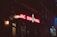 A dimly lit street scene features a neon sign with the words 'JAC SON HOLE' in bright red letters, accompanied by a glowing silhouette of a cowboy riding a bucking bronco. The storefront is dark, with subtle lighting highlighting the brick wall and an entrance. The surrounding area appears to have closed shops and is relatively quiet.