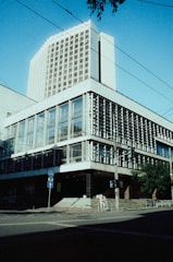 A modernist architectural building with large glass windows and concrete details. A taller structure rises in the background, showcasing rectangular windows. The scene includes pedestrian pathways, traffic signs, and a few people walking, likely in an urban setting.