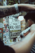 A person is getting a haircut in a space decorated with license plates, stickers, and graffiti. Texas and Dominican Republic license plates are visible on a brick wall. A tattooed arm and a barber's clipper are in the foreground.