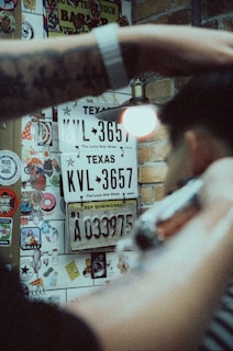 A person is getting a haircut in a space decorated with license plates, stickers, and graffiti. Texas and Dominican Republic license plates are visible on a brick wall. A tattooed arm and a barber's clipper are in the foreground.