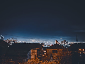 A nighttime cityscape featuring a mixture of illuminated buildings and darkened rooftops in the foreground. The sky is a deep navy blue, with city lights twinkling below, providing a contrast to the dark silhouettes of structures. An electrical tower stands out to the right side, adding an industrial element to the scene.
