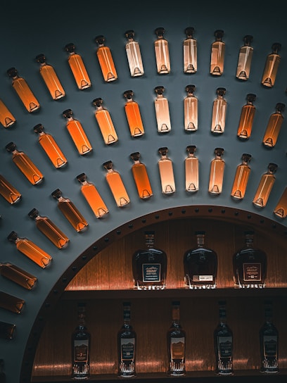 A display of glass bottles filled with amber-colored liquid, arranged in an arc on a dark wall. Below, larger bottles of spirits, likely whiskey or brandy, are showcased on shelves with warm lighting.