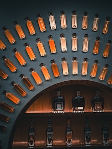 A display of glass bottles filled with amber-colored liquid, arranged in an arc on a dark wall. Below, larger bottles of spirits, likely whiskey or brandy, are showcased on shelves with warm lighting.