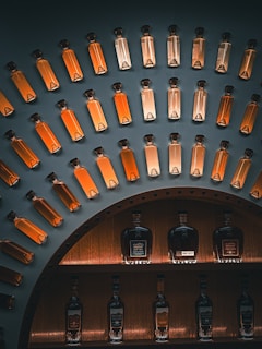 A display of glass bottles filled with amber-colored liquid, arranged in an arc on a dark wall. Below, larger bottles of spirits, likely whiskey or brandy, are showcased on shelves with warm lighting.