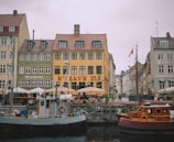 A picturesque waterfront scene featuring colorful historic buildings with red-tiled roofs lining a canal. Several boats are docked in the foreground, including both a wooden and metal vessel. People are sitting under umbrellas at outdoor cafes along the quay, creating a lively yet relaxed atmosphere. Danish flags can be seen flying from some of the boats.