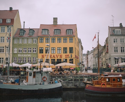 A picturesque waterfront scene featuring colorful historic buildings with red-tiled roofs lining a canal. Several boats are docked in the foreground, including both a wooden and metal vessel. People are sitting under umbrellas at outdoor cafes along the quay, creating a lively yet relaxed atmosphere. Danish flags can be seen flying from some of the boats.