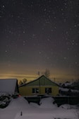 A starry night sky over a residential area with a yellow house in the foreground. The house is surrounded by a snow-covered landscape, and the light pollution creates a subtle glow on the horizon.