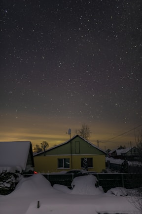 A starry night sky over a residential area with a yellow house in the foreground. The house is surrounded by a snow-covered landscape, and the light pollution creates a subtle glow on the horizon.