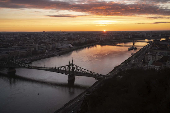 A panoramic view of the Howrah Bridge at sunset, glowing warmly over the Hooghly River.