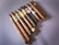 A collection of ten cigars is neatly arranged on a flat surface, each wrapped in different colored bands that provide branding and flavor information. The cigars vary slightly in color from light to dark brown, suggesting different tobacco types and origins. The surface they rest on is smooth and softly shadowed.