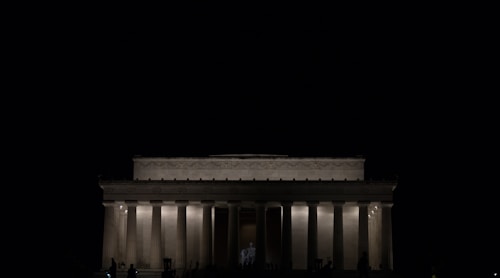 A large, classical-style building with tall columns is illuminated against a dark background. The lighting highlights its architectural features, giving it a majestic appearance. The image conveys a sense of grandeur and solemnity.