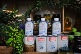 Several cans of extra virgin olive oil are displayed alongside various food items, including jars, canned goods, and green leafy plants. The olive oil cans are prominently placed in the center and are labeled 'Lindsay Naturals' and 'La Tourangelle' with colorful and vibrant labels.