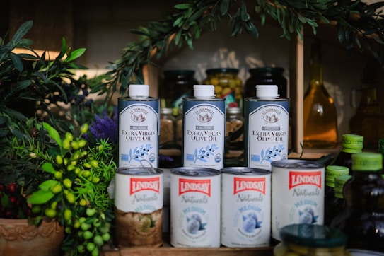 Several cans of extra virgin olive oil are displayed alongside various food items, including jars, canned goods, and green leafy plants. The olive oil cans are prominently placed in the center and are labeled 'Lindsay Naturals' and 'La Tourangelle' with colorful and vibrant labels.