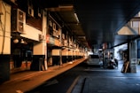 A dimly lit alleyway displays a row of air conditioning units mounted on the walls above, with a parked white car and a small utility vehicle nearby. The area appears to be part of a commercial marketplace with various signs written in Korean, suggesting a business district.