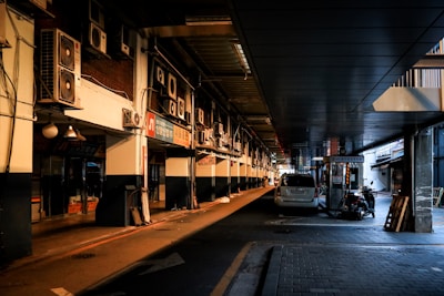 A dimly lit alleyway displays a row of air conditioning units mounted on the walls above, with a parked white car and a small utility vehicle nearby. The area appears to be part of a commercial marketplace with various signs written in Korean, suggesting a business district.