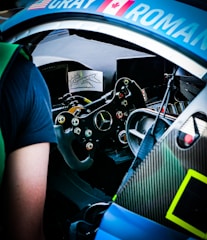 Inside view of a sports car cockpit showcasing a sophisticated steering wheel with numerous buttons, a dashboard screen, and a partial view of the driver's seat. The car seems to be equipped for racing with various controls and a track map displayed.