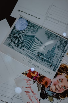 A collection of vintage postcards is displayed. One postcard features an illustration of an elegant building surrounded by winter scenery with trees. Another postcard displays a smiling child, possibly celebrating a festive occasion. The text on the postcards is written in Russian.