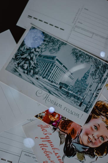 A collection of vintage postcards is displayed. One postcard features an illustration of an elegant building surrounded by winter scenery with trees. Another postcard displays a smiling child, possibly celebrating a festive occasion. The text on the postcards is written in Russian.