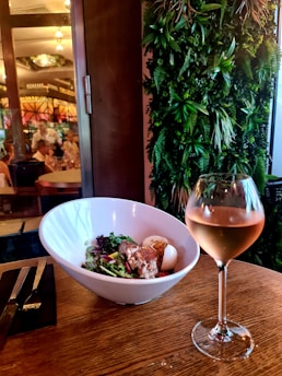 A bowl of salad with assorted greens, a boiled egg, and grilled chicken sits on a wooden table. Next to it is a glass of rosé wine. The background features a vertical garden with lush green plants and a restaurant interior with diners visible through a glass door.