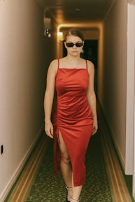A person wearing a red satin dress and sunglasses walks confidently down a hotel hallway with soft lighting and intricate carpet design.