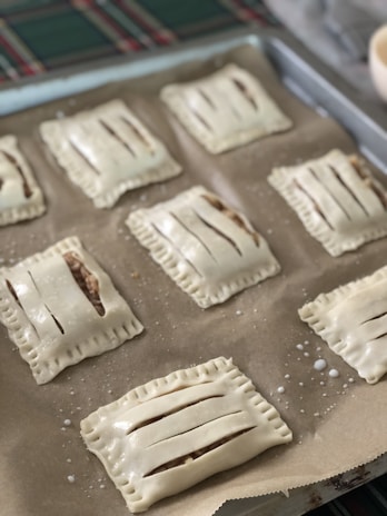 Several rectangular pastries with slits on top are arranged on a baking sheet covered with parchment paper. The dough appears unbaked, and the pastries are evenly spaced on the tray, ready to be baked. A green and red checkered cloth is partially visible in the background.