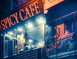 A nighttime view of a modern cafe with a glowing sign that reads 'Spicy Cafe.' The interior features warm lighting and framed artwork on the walls. A person is seen inside, leaning over a counter. Outside, there is a weathered building facade with exposed pipes and wires.