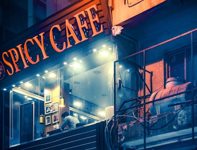 A nighttime view of a modern cafe with a glowing sign that reads 'Spicy Cafe.' The interior features warm lighting and framed artwork on the walls. A person is seen inside, leaning over a counter. Outside, there is a weathered building facade with exposed pipes and wires.