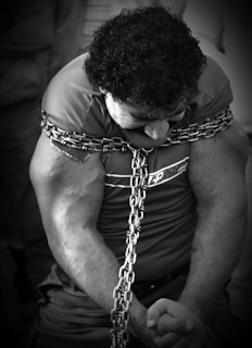 A person with muscular arms is bound tightly with thick metal chains around the shoulders and torso. The individual is looking downward, and their expression is intense, suggesting strain or concentration.