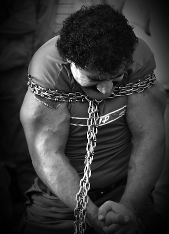 A person with muscular arms is bound tightly with thick metal chains around the shoulders and torso. The individual is looking downward, and their expression is intense, suggesting strain or concentration.