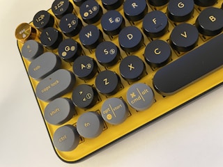 A close-up view of a mechanical keyboard with retro-style round keys. The keys are a mix of dark gray and gold with yellow accents, creating a unique and vibrant aesthetic. The keyboard layout includes standard keys like 'tab', 'caps lock', 'shift', 'ctrl', and more. Notably, the 'esc' key stands out in gold.