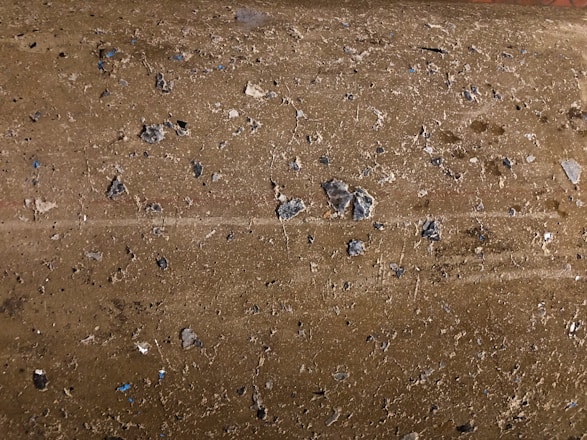 A close-up view of a rough, textured concrete surface with small pebbles and stones embedded in it. The surface has an uneven appearance with various shades of gray and brown. Small bits of blue can be seen among the natural tones.