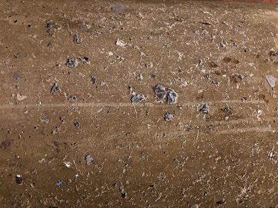 A close-up view of a rough, textured concrete surface with small pebbles and stones embedded in it. The surface has an uneven appearance with various shades of gray and brown. Small bits of blue can be seen among the natural tones.