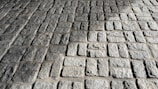 A close-up view of a stone pavement consisting of rectangular cobblestones arranged in a regular pattern. The uneven texture of the stones is highlighted by the sunlight, creating shadows and emphasizing their rugged surface.