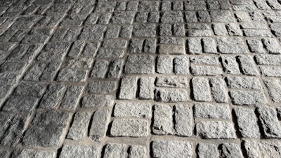 A close-up view of a stone pavement consisting of rectangular cobblestones arranged in a regular pattern. The uneven texture of the stones is highlighted by the sunlight, creating shadows and emphasizing their rugged surface.