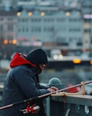 A person is concentrating on preparing fishing gear. The individual is dressed warmly in a blue coat with a red hood, possibly due to cold weather. The scene is set on a bridge or platform against a blurred urban background, with bokeh lights indicating a city setting.