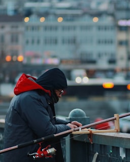 A person is concentrating on preparing fishing gear. The individual is dressed warmly in a blue coat with a red hood, possibly due to cold weather. The scene is set on a bridge or platform against a blurred urban background, with bokeh lights indicating a city setting.