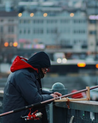 A person is concentrating on preparing fishing gear. The individual is dressed warmly in a blue coat with a red hood, possibly due to cold weather. The scene is set on a bridge or platform against a blurred urban background, with bokeh lights indicating a city setting.