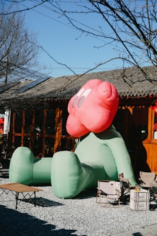 A large, inflatable sculpture with a pink head and green body sits in an outdoor setting on gravel. The sculpture resembles a cartoonish figure with large, round eyes. It is positioned in front of a wooden structure, likely a building, with clear skies overhead and bare tree branches in the background. There are folding chairs and a small table nearby, creating a relaxed outdoor area.