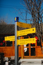A signpost with four yellow directional signs indicating locations such as Toilet, Smoking Corner, Picture Corner, and Vending Machine. The signs are written in both English and Chinese. The background includes a traditional-style building with wooden panels, red circular decorations, and bare tree branches.