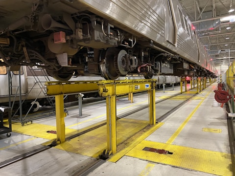 A large train is elevated on yellow metal platforms inside a maintenance workshop. The undercarriage is visible with various mechanical components and wheels. The setting is industrial with high ceilings and bright overhead lights.