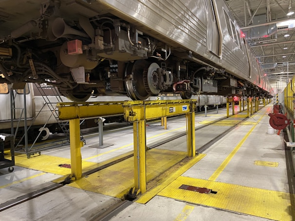 A large train is elevated on yellow metal platforms inside a maintenance workshop. The undercarriage is visible with various mechanical components and wheels. The setting is industrial with high ceilings and bright overhead lights.
