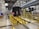 A metro train is positioned inside a maintenance facility. The train is elevated above the ground, supported by a track system, allowing for inspection or repairs. Various equipment and tools are visible around the area, with bright lighting illuminating the space. A worker is seen in the background, suggesting an active maintenance process.