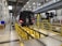 A metro train is positioned inside a maintenance facility. The train is elevated above the ground, supported by a track system, allowing for inspection or repairs. Various equipment and tools are visible around the area, with bright lighting illuminating the space. A worker is seen in the background, suggesting an active maintenance process.